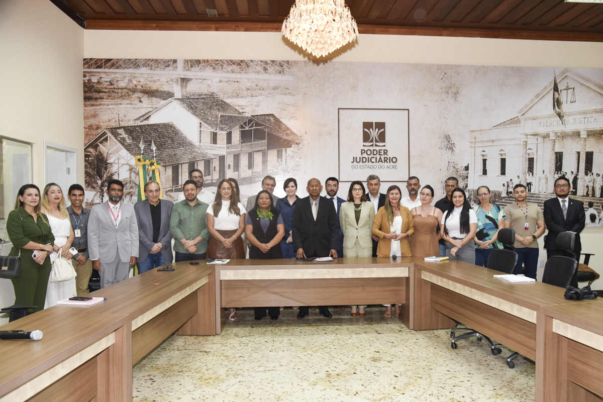 A imagem mostra um grupo de aproximadamente 25 pessoas reunidas em uma sala de reuniões do Poder Judiciário do Estado do Acre. Elas estão posicionadas em pé, lado a lado, formando uma linha curva atrás de uma mesa em formato de "U", indicando que a foto foi tirada ao final de um evento ou reunião formal. No centro da composição, está um homem negro de terno escuro, sugerindo posição de destaque ou liderança na ocasião. Ao lado dele, está uma mulher indígena com colares e adereços de folhas, sinalizando a presença de representatividade indígena no evento. À esquerda da imagem, estão as bandeiras do Brasil e do Acre. Ao fundo, um grande painel fotográfico exibe imagens históricas de prédios e ruas da região, incluindo o Palácio da Justiça, reforçando o ambiente institucional. No centro do painel está o logotipo do Poder Judiciário do Estado do Acre. O local tem iluminação clara e um lustre pendente no teto, reforçando o caráter solene da reunião. Trata-se de um evento institucional com diversidade de participantes — servidores públicos, representantes indígenas e autoridades —, indicando um momento de diálogo ou articulação entre diferentes setores sociais.