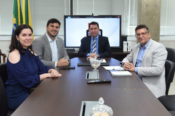 Os indivíduos presentes na imagem são quatro adultos. Três homens e uma mulher, todos vestidos com roupas de negócios formais. Os participantes estão sentados em cadeiras de escritório, com os braços apoiando-se sobre a mesa. Todos os olhares dos participantes estão voltados para a frente. A mulher está vestindo uma camisa azul com os ombros à mostra e, do lado esquerdo da mesa, o homem ao seu lado está vestindo um terno cinza com camisa social branca. Na ponta da mesa está um outro homem vestindo um terno preto, camisa social azul claro e gravata listrada. Do lado direito da mesa, se encontra um homem vestindo um terno cinza com uma camisa social azul claro com listras brancas.