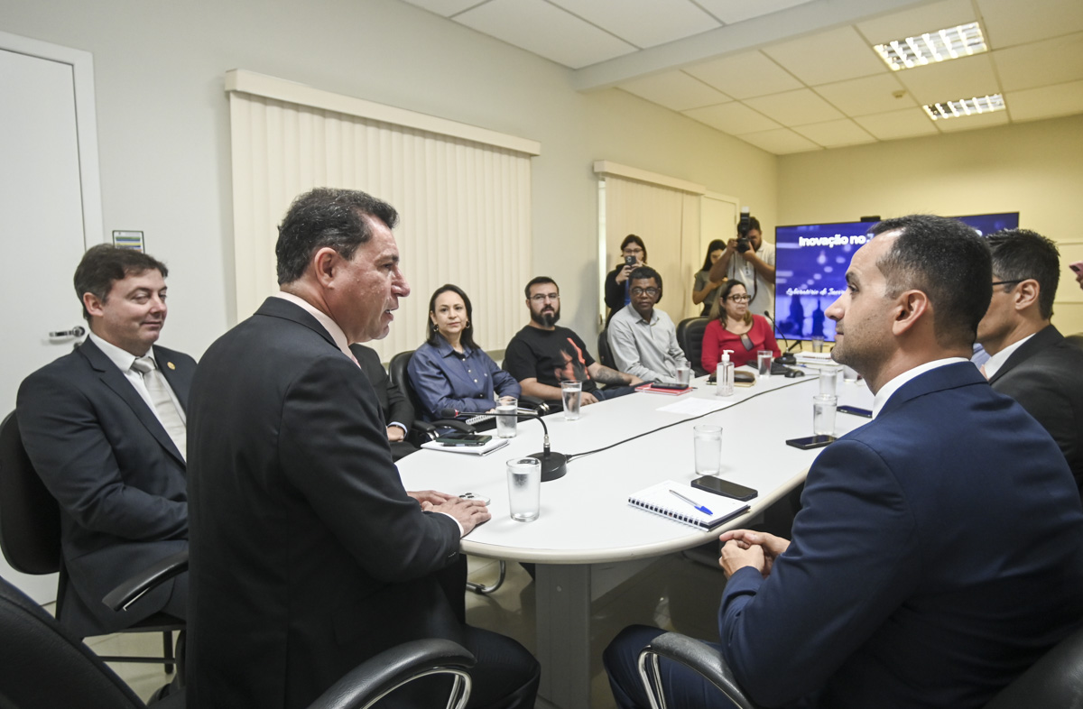 Na imagem, oito pessoas estão reunidos ao redor de uma mesa oval, todos vestem roupas formais e aparentam está concentrados em um assunto importante