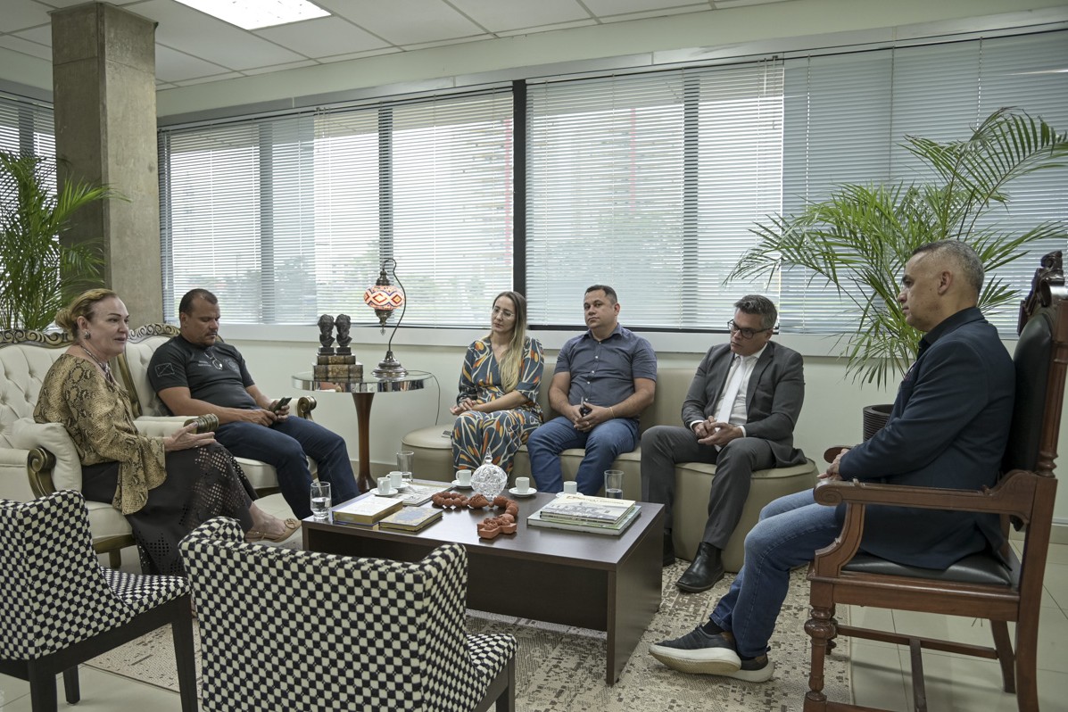 A imagem mostra seis pessoas onde são duas mulheres e quatro homens reunidas em uma sala de reuniões ao redor de uma pequena mesa de madeira. Todos parecem estar participando de uma reunião com copo de água, xicara de café e ambos mexendo no celular. A sala tem um design corporativo, as pessoas na foto estão sorrindo e parecem engajadas na reunião. A iluminação é clara, vinda das luzes no teto.