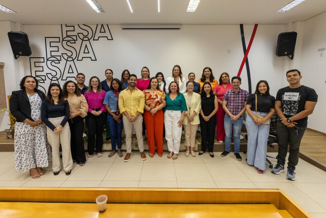A imagem mostra um grupo de aproximadamente 25 pessoas posando para uma foto coletiva em um ambiente interno, provavelmente uma sala de eventos ou auditório. Todos estão de pé, formando duas fileiras — a da frente com pessoas em pé diretamente no chão e a de trás sobre uma pequena plataforma. A maioria das pessoas está sorrindo, indicando um clima descontraído e positivo. No fundo da imagem, há a sigla "ESA" (repetida várias vezes na parede), que pode indicar que o local é ligado à Escola Superior da Advocacia ou alguma outra instituição educacional ou profissional com a mesma sigla. As paredes são brancas com detalhes decorativos em vermelho e azul escuro. Há caixas de som nas extremidades superiores da sala, reforçando a ideia de um ambiente preparado para eventos ou apresentações. As roupas das pessoas variam entre estilos casuais e semiformal, com uma diversidade de cores vibrantes, o que contribui para uma atmosfera alegre e acolhedora. No canto inferior da imagem, vê-se parcialmente uma bancada de madeira e um copo descartável, possivelmente de café, o que indica que o evento pode incluir momentos de confraternização. Parece tratar-se de um encontro, curso, palestra ou formatura promovida por uma instituição de ensino ou formação profissional.