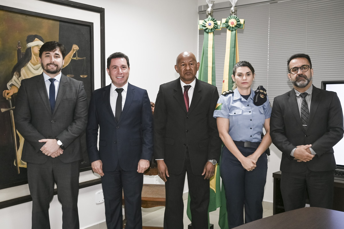 A imagem mostra cinco pessoas posando formalmente para uma foto em um ambiente institucional. Todos estão vestidos com trajes sociais ou uniformes, e o cenário reforça o tom oficial do encontro. Da esquerda para a direita: Um homem de terno escuro com camisa branca e gravata azul-marinho. Um homem ao centro também de terno escuro, gravata escura e camisa branca, sorrindo. Um homem de pele negra, com expressão séria, vestindo terno preto e gravata bordô. Uma mulher fardada, provavelmente de uma corporação policial ou militar, com insígnias no ombro e a bandeira do Acre no uniforme. Um homem de terno escuro, camisa cinza e gravata com listras diagonais, com as mãos cruzadas à frente. Ao fundo, aparecem duas bandeiras do Brasil com laços verde-amarelos e uma pintura que retrata a figura da justiça (mulher vendada segurando a balança e uma espada). A composição e os trajes sugerem uma reunião ou visita de caráter oficial entre representantes do poder público e das forças de segurança.