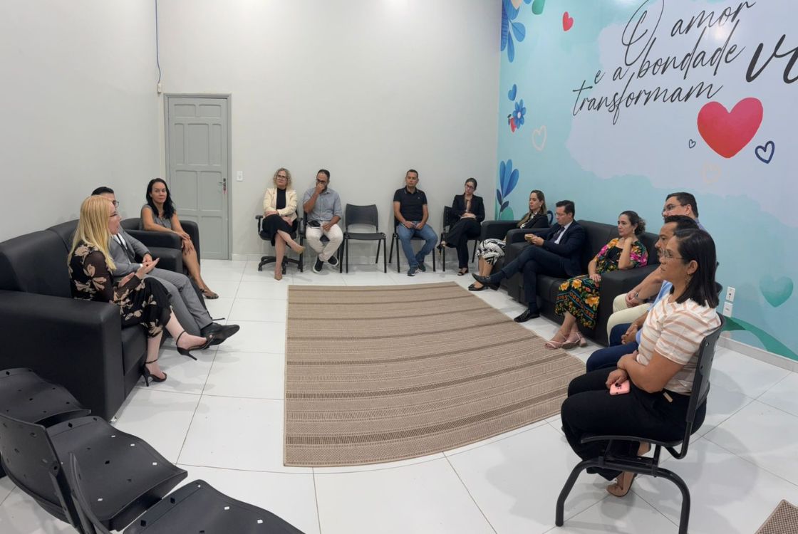 A imagem mostra um grupo de aproximadamente 16 pessoas participando de uma reunião em um ambiente informal e acolhedor. Os participantes estão sentados em sofás e cadeiras dispostas em círculo, sugerindo um formato de roda de conversa. A parede ao fundo é decorada com desenhos de corações, flores e a frase: “O amor e a bondade transformam vidas”, o que reforça o clima de empatia e diálogo. As pessoas aparentam estar concentradas, ouvindo uma mulher que fala, sentada à esquerda da imagem. A reunião parece tratar de temas sociais ou institucionais, com a presença de servidores públicos, profissionais de diversas áreas e autoridades. O ambiente transmite acolhimento, integração e escuta ativa.