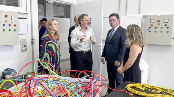 A imagem mostra um grupo de pessoas em um ambiente que parece ser um laboratório ou sala técnica, possivelmente de uma instituição de ensino ou centro de pesquisa. No local, há painéis de controle com botões e luzes indicadoras nas paredes e fios elétricos coloridos (vermelhos, verdes, amarelos e azuis) organizados sobre uma mesa, o que sugere atividades relacionadas à eletricidade, automação ou engenharia. No centro da imagem, um homem de camisa xadrez clara gesticula com as mãos enquanto aparentemente explica algo aos demais. Ao seu redor estão quatro pessoas: duas mulheres (uma de vestido preto e outra com uma roupa estampada), e dois homens (um em traje formal, terno e gravata, e outro ao fundo). Todos parecem atentos à explicação, e o clima geral é de visita técnica ou apresentação institucional. Há também uma placa na porta com o texto parcialmente visível “…tricos”, indicando que pode ser um laboratório de “circuitos elétricos” ou algo semelhante.