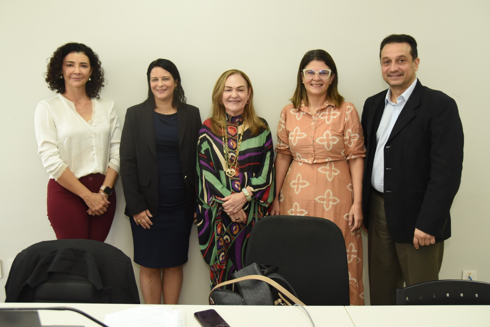 A imagem mostra cinco pessoas em pé, alinhadas lado a lado, posando para uma foto em um ambiente corporativo ou institucional. A parede de fundo é lisa, de cor clara, e à frente há uma mesa com alguns itens visíveis, como uma mochila, um caderno espiral, um celular e fios de equipamentos eletrônicos. As pessoas estão bem vestidas, com roupas sociais e profissionais, demonstrando um clima formal. No centro está uma mulher com um vestido colorido de estampas geométricas e um colar dourado chamativo, que aparece também em outras fotos, sugerindo que pode ocupar uma posição de destaque, como convidada ou autoridade. As outras pessoas, duas mulheres à esquerda e uma mulher e um homem à direita, também demonstram postura respeitosa e cordial, o que reforça a ideia de um encontro institucional ou reunião importante.