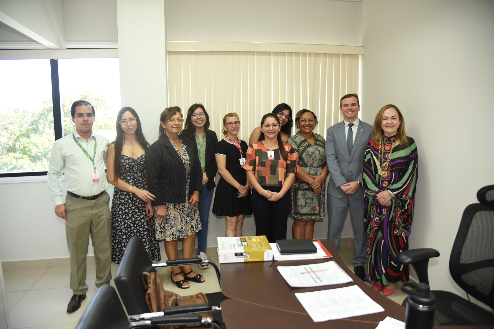 A imagem mostra um grupo de treze pessoas reunidas em um escritório institucional para uma foto coletiva. Todos estão em pé, lado a lado, em frente a uma persiana vertical branca. À esquerda, há uma grande janela com vista para árvores, sugerindo que a sala está em um prédio alto. Em primeiro plano, aparecem parcialmente cadeiras e uma mesa com documentos, livros e uma mochila, indicando um ambiente de trabalho ativo. As pessoas estão vestidas de forma variada, com trajes que vão desde o social completo (como terno e vestido formal) até roupas mais casuais com crachás, o que reforça o caráter funcional e colaborativo da ocasião. O homem de terno cinza claro e a mulher com vestido colorido de estampas geométricas (presente nas imagens anteriores) estão próximos ao centro, sugerindo que podem ser figuras centrais no evento. O clima da imagem é de formalidade e cordialidade, indicando provavelmente uma visita institucional, reunião de trabalho, ou registro de um momento relevante dentro de um órgão público ou organização.