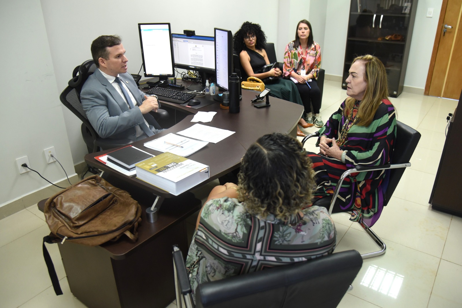 A imagem mostra uma reunião formal em um ambiente de escritório. Um homem, vestido com terno cinza e gravata, está sentado atrás de uma mesa de trabalho, aparentemente conduzindo a conversa. Ele está interagindo com quatro mulheres, duas sentadas à sua frente e duas ao fundo, que parecem estar acompanhando ou participando da reunião. Na mesa, há dois monitores de computador, livros (incluindo um intitulado “Manual de Direito Penal”), cadernos, papéis, uma mochila de couro, uma garrafa térmica e outros itens de escritório. As mulheres estão bem-vestidas, sugerindo um encontro profissional. A sala tem aparência moderna e organizada, com piso claro e paredes neutras. A reunião parece estar ocorrendo em um ambiente institucional, possivelmente ligado ao setor jurídico, educacional ou administrativo.