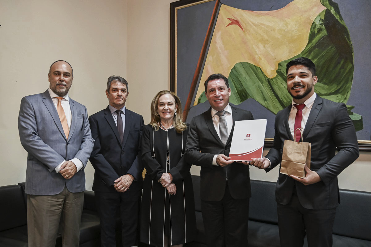 Quatro pessoas posam para uma foto em um ambiente que remete a uma cerimônia institucional ou reunião formal. Entre elas, há uma mulher e três homens. Dois dos homens estão vestidos com ternos pretos, enquanto o terceiro veste um terno cinza-claro com calça bege. Um dos homens de terno preto segura um documento, sugerindo a entrega ou assinatura oficial de algo, enquanto o homem à esquerda segura um saco de presente, reforçando o clima de celebração. A composição e o ambiente indicam uma ocasião formal, possivelmente uma cerimônia de entrega ou reconhecimento institucional.