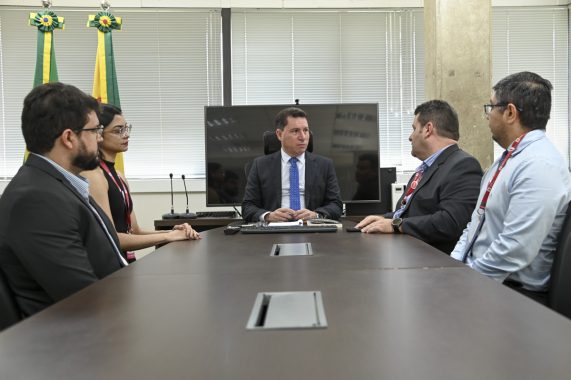 A imagem mostra seis pessoas — quatro homens e duas mulheres — sentadas ou posicionadas ao redor de uma mesa de reunião em um ambiente institucional. Ao fundo, há duas bandeiras do estado do Acre, o que sugere que a reunião ocorreu em uma repartição pública estadual. No centro da imagem, sentado à cabeceira da mesa, está um homem de terno escuro e gravata azul, que parece ocupar o cargo de maior autoridade no encontro. Atrás dele, há um monitor e microfones, indicando um espaço preparado para reuniões oficiais ou videoconferências. Todos estão vestidos com roupas formais e usam crachás de identificação, o que reforça o caráter corporativo ou governamental da reunião. A expressão dos participantes é cordial, sugerindo um encontro produtivo e colaborativo.