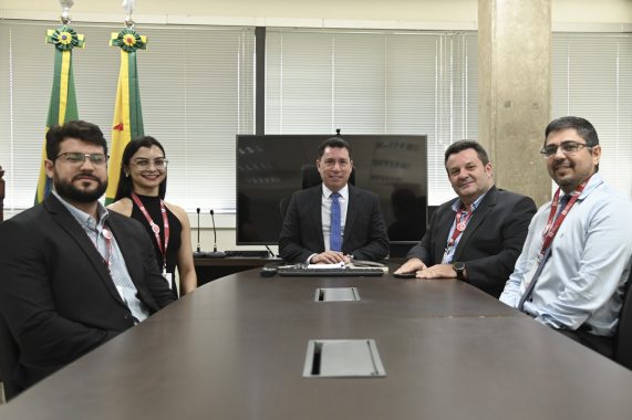 A imagem mostra seis pessoas — quatro homens e duas mulheres — sentadas ou posicionadas ao redor de uma mesa de reunião em um ambiente institucional. Ao fundo, há duas bandeiras do estado do Acre, o que sugere que a reunião ocorreu em uma repartição pública estadual. No centro da imagem, sentado à cabeceira da mesa, está um homem de terno escuro e gravata azul, que parece ocupar o cargo de maior autoridade no encontro. Atrás dele, há um monitor e microfones, indicando um espaço preparado para reuniões oficiais ou videoconferências. Todos estão vestidos com roupas formais e usam crachás de identificação, o que reforça o caráter corporativo ou governamental da reunião. A expressão dos participantes é cordial, sugerindo um encontro produtivo e colaborativo.