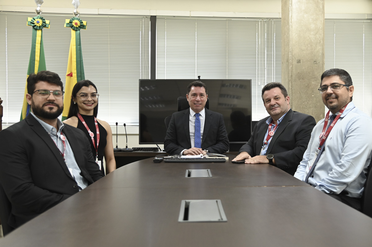 A imagem mostra seis pessoas — quatro homens e duas mulheres — sentadas ou posicionadas ao redor de uma mesa de reunião em um ambiente institucional. Ao fundo, há duas bandeiras do estado do Acre, o que sugere que a reunião ocorreu em uma repartição pública estadual. No centro da imagem, sentado à cabeceira da mesa, está um homem de terno escuro e gravata azul, que parece ocupar o cargo de maior autoridade no encontro. Atrás dele, há um monitor e microfones, indicando um espaço preparado para reuniões oficiais ou videoconferências. Todos estão vestidos com roupas formais e usam crachás de identificação, o que reforça o caráter corporativo ou governamental da reunião. A expressão dos participantes é cordial, sugerindo um encontro produtivo e colaborativo.