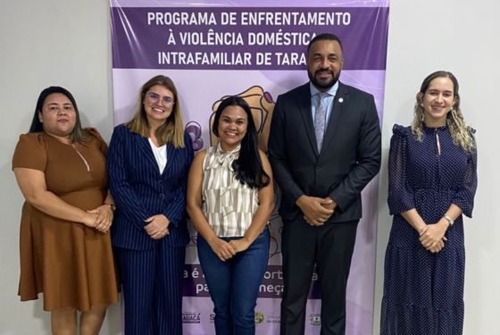 A imagem mostra cinco pessoas posando para uma foto em frente a uma faixa institucional do “Programa de Enfrentamento à Violência Doméstica e Intrafamiliar de Tarauacá”. Este parece ser um momento formal, possivelmente o registro de uma equipe envolvida na organização ou execução do programa. Detalhes da imagem: Pessoas na imagem: Três mulheres à esquerda e uma mulher à direita usam roupas sociais — vestidos ou ternos, indicando um contexto profissional. Um homem, ao centro-direita, veste terno e gravata, sugerindo um cargo de liderança ou representação institucional (possivelmente juiz, promotor ou gestor público). Todos estão sorrindo, demonstrando um momento de cordialidade e formalidade. Faixa ao fundo: Tem fundo roxo e traz a logomarca do programa e parceiros institucionais (embora não estejam completamente visíveis). Além do título do programa, há um símbolo de duas mãos segurando uma figura feminina, representando apoio e proteção à mulher. Interpretação: A imagem retrata uma equipe técnica e institucional diretamente envolvida com ações de enfrentamento à violência doméstica e intrafamiliar no município de Tarauacá (AC). O cenário é típico de eventos públicos, ações de conscientização ou lançamento de projetos sociais. A disposição e o vestuário dos presentes reforçam o compromisso profissional e o caráter oficial da ocasião.