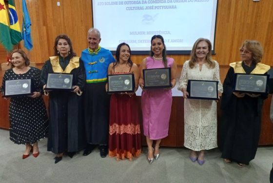 A imagem mostra um grupo de sete pessoas posando para uma foto oficial em um ambiente solene, com parede de madeira e bandeiras ao fundo (incluindo a do Brasil). Eles estão em frente a um telão com a seguinte inscrição visível: "Ato solene de outorga da Comenda da Ordem do Mérito Cultural José Potyguara" "Mulher: resistência, coragem, determinação e ação" A ocasião é claramente uma cerimônia de homenagem, voltada à valorização de mulheres que se destacam em suas áreas de atuação. Entre os homenageados, cinco mulheres seguram placas comemorativas de homenagem. Algumas delas estão vestidas com becas cerimoniais com detalhes dourados nos ombros. Duas mulheres, ao centro, estão vestidas com roupas sociais elegantes em tons de vermelho e rosa. Um homem, também homenageado ou integrante da organização, veste uma beca azul com detalhes verdes e dourados. Todos sorriem, demonstrando orgulho e alegria pelo momento de reconhecimento, o que reforça o caráter celebrativo e institucional do evento.