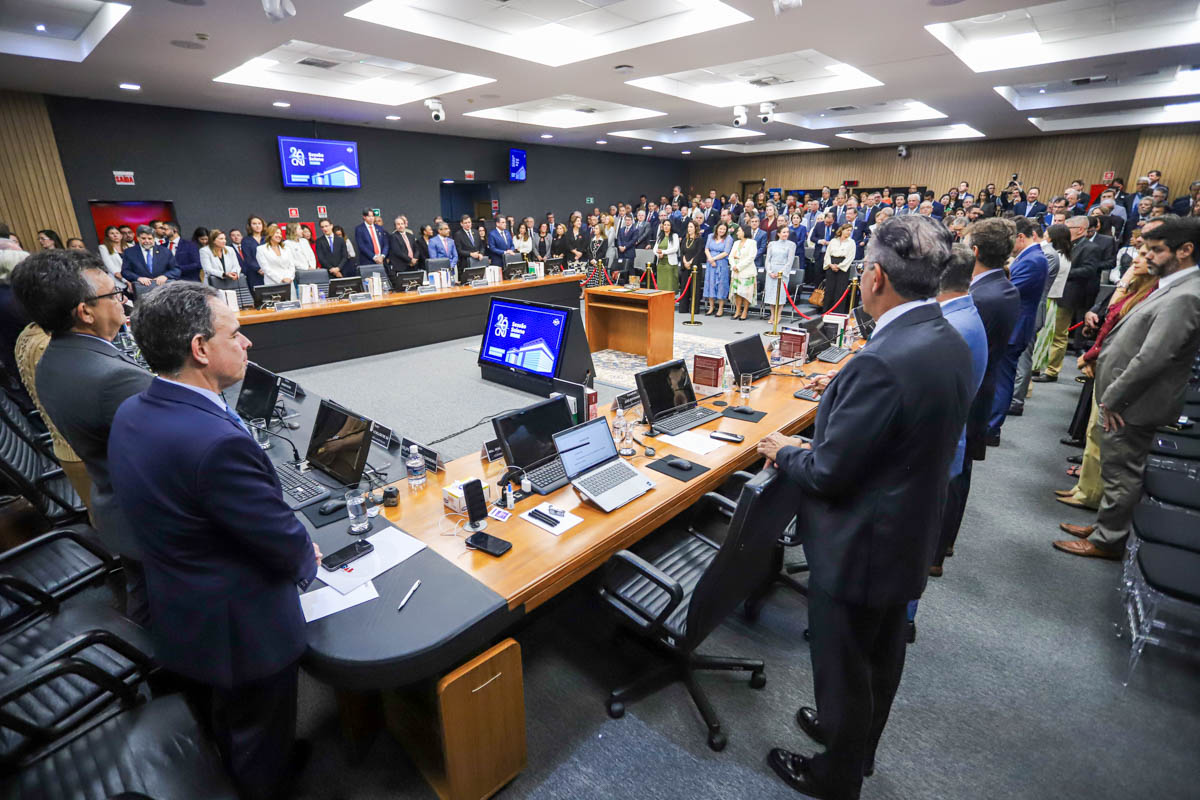 A imagem mostra uma sessão solene ou uma reunião formal em um auditório moderno, provavelmente pertencente a uma instituição governamental, jurídica ou corporativa de alto nível. A cena retrata: Um salão com um grande número de pessoas em pé, sugerindo o início ou encerramento do evento, ou um momento solene como o hino nacional. Na frente, há uma mesa em formato de “U” com vários notebooks abertos, microfones, garrafas de água e placas de identificação, indicando que esse espaço é reservado para os participantes principais ou autoridades. Um púlpito ao centro, sobre um tapete, sugere que discursos ou apresentações estão ocorrendo ou irão ocorrer. Ao fundo, há muitas pessoas organizadas em pé, bem-vestidas, sugerindo um público seleto de convidados, funcionários ou membros de uma organização. Telas ao redor da sala exibem um logotipo e a mensagem “26 anos”, indicando que o evento celebra o aniversário de 26 anos de alguma entidade ou instituição. O ambiente é bem iluminado, moderno e transmite uma atmosfera de formalidade, profissionalismo e respeito institucional.