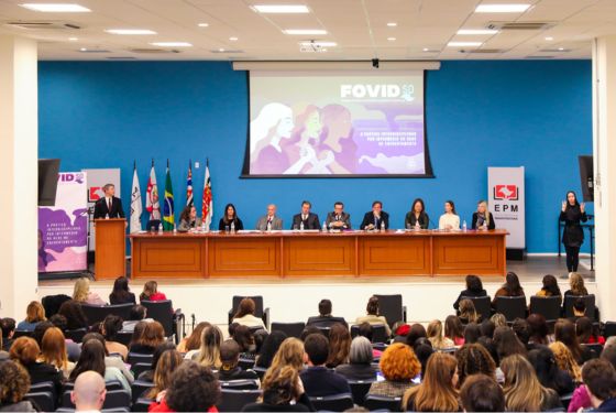A imagem mostra um auditório durante um evento formal. Na parte frontal, há uma mesa ampla com cerca de dez pessoas sentadas, provavelmente autoridades ou palestrantes. Um dos participantes está em pé, discursando de um púlpito à esquerda. Ao fundo, há um telão com a projeção de uma imagem que contém o título “FOVID 50” e ilustrações estilizadas de mulheres, sugerindo que o evento está relacionado a temas de gênero ou violência doméstica, como indica o subtítulo parcialmente visível. À esquerda do telão, estão posicionadas várias bandeiras — possivelmente de estados brasileiros ou entidades oficiais — e banners institucionais. À direita, há uma intérprete de Libras (Língua Brasileira de Sinais), sinalizando acessibilidade para pessoas surdas. O auditório está cheio, com o público sentado e voltado para o palco, evidenciando um evento de grande participação. O ambiente é profissional e bem iluminado, sugerindo um seminário, fórum ou conferência institucional.