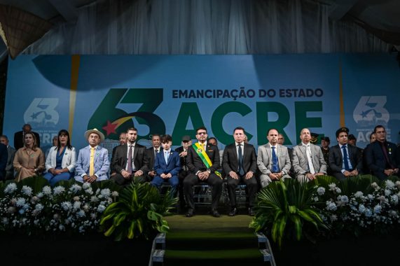 A imagem mostra uma solene cerimônia cívica, em comemoração aos 63 anos da emancipação do Estado do Acre, como indicado pelo grande painel de fundo com os dizeres "63 anos - Emancipação do Estado do Acre". Elementos centrais da imagem: Ao centro da primeira fileira de autoridades, está o governador do Acre, vestindo um terno preto com faixa oficial verde, amarela e vermelha, além de um broche ou medalha oficial no peito. À sua esquerda (na imagem), há um menino de terno azul claro, aparentando ser um convidado especial, filho ou representante simbólico da juventude, também em posição de destaque. Ao lado do menino e nas demais cadeiras da primeira fileira estão diversas autoridades: Homens de terno escuro e claro, alguns com estrela verde no peito (símbolo usado por alguns dos presentes nas imagens anteriores, provavelmente indicando condecoração ou cargo). Todos estão sentados em postura formal, com semblantes sérios ou neutros. Ao fundo, observa-se o logotipo comemorativo com o número 63 e a bandeira do Acre estilizada. A frente do palco está decorada com arranjos de folhas verdes e flores brancas, realçando o tom cerimonial do evento. Interpretação: A imagem transmite um momento de respeito e solenidade, marcando a celebração de um marco importante na história política do Acre. A presença do governador, de representantes civis, militares e de diferentes gerações sugere um evento plural e institucional, com forte valor simbólico para o estado.