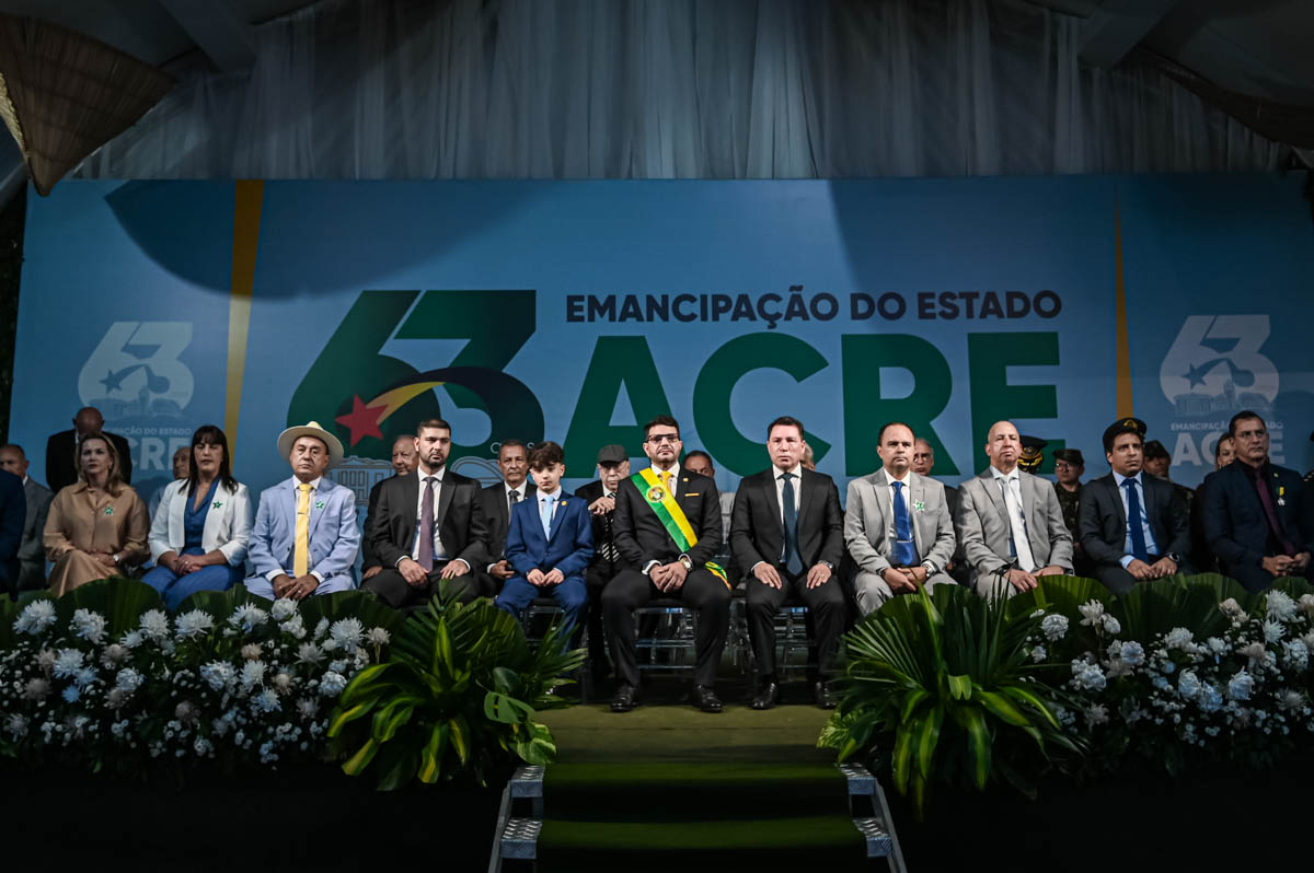 A imagem mostra uma solene cerimônia cívica, em comemoração aos 63 anos da emancipação do Estado do Acre, como indicado pelo grande painel de fundo com os dizeres "63 anos - Emancipação do Estado do Acre". Elementos centrais da imagem: Ao centro da primeira fileira de autoridades, está o governador do Acre, vestindo um terno preto com faixa oficial verde, amarela e vermelha, além de um broche ou medalha oficial no peito. À sua esquerda (na imagem), há um menino de terno azul claro, aparentando ser um convidado especial, filho ou representante simbólico da juventude, também em posição de destaque. Ao lado do menino e nas demais cadeiras da primeira fileira estão diversas autoridades: Homens de terno escuro e claro, alguns com estrela verde no peito (símbolo usado por alguns dos presentes nas imagens anteriores, provavelmente indicando condecoração ou cargo). Todos estão sentados em postura formal, com semblantes sérios ou neutros. Ao fundo, observa-se o logotipo comemorativo com o número 63 e a bandeira do Acre estilizada. A frente do palco está decorada com arranjos de folhas verdes e flores brancas, realçando o tom cerimonial do evento. Interpretação: A imagem transmite um momento de respeito e solenidade, marcando a celebração de um marco importante na história política do Acre. A presença do governador, de representantes civis, militares e de diferentes gerações sugere um evento plural e institucional, com forte valor simbólico para o estado.