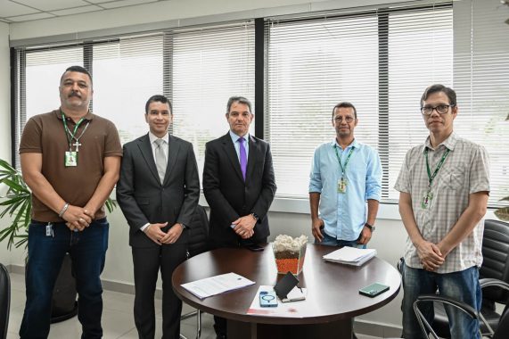 A imagem mostra cinco homens posando para uma foto em um ambiente corporativo, provavelmente uma sala de reuniões. Três deles (o primeiro da esquerda, o quarto e o quinto da esquerda para a direita) estão usando crachás com cordões verdes, sugerindo que sejam funcionários da mesma instituição. Dois deles (o segundo e o terceiro da esquerda) estão trajando ternos formais, o que pode indicar que são visitantes ou ocupam cargos de liderança. Ao centro, há uma mesa redonda com alguns documentos, um vaso decorativo com pedras e grãos, um crachá, um celular e um caderno. As janelas ao fundo possuem persianas horizontais parcialmente fechadas, e o ambiente é bem iluminado por luz natural. A atmosfera é de formalidade e profissionalismo, típica de uma reunião ou assinatura de acordos institucionais.