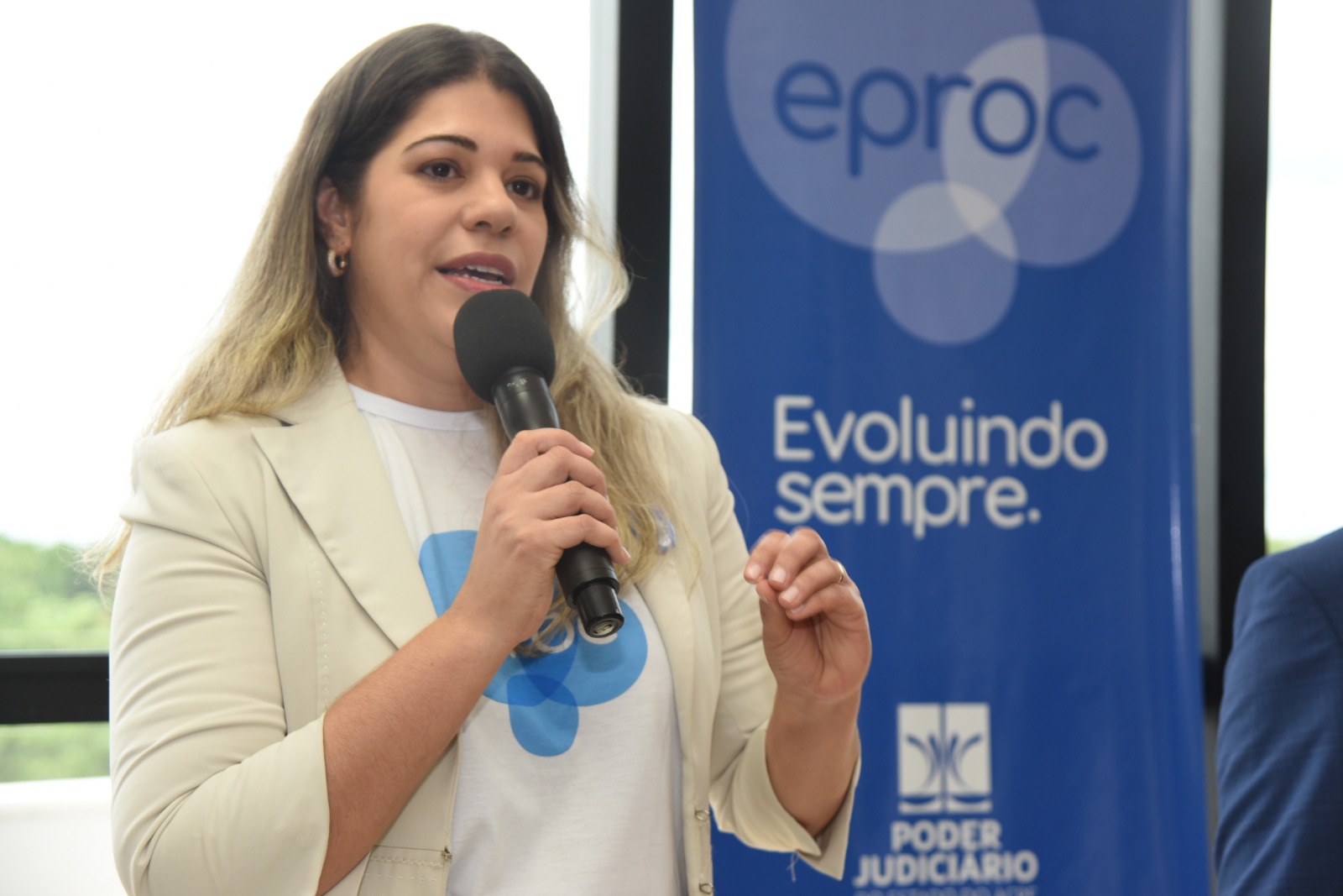 Juíza Louise. É loira. Está de pé segurando um microfone. Veste uma blusa branca com a logo do eproc, que é um cículo azul. Ela veste um blazer de cor nude. Ao fundo da foto tem  um banner azul com a logo do eproc